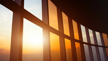 Im Vordergrund Fensterfront und Decke eines hohen Raumes. Manche Fensterscheiben sind getönt. Im Hintergrund der Himmel mit durchs Fenster scheinender Sonne.