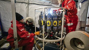 Zwei Forscherinnen stehen bzw. sitzen neben einem Messinstrument in einem Zelt. Das Messinstrument besteht unter anderem aus mehreren gasflaschenähnlichen Behältern.