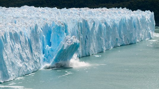 Das Foto zeigt den Perito-Moreno-Gletscher. Eine Hälfte des Bildes zeigt den Gletscher mit seiner Front, von der ein Stück abbricht. In der anderen Hälfte ist Wasser. 