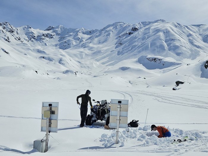 Ein Mensch in einer verschneiten Landschaft steht neben einem Schneemobil, auf dem eine technische Apparatur angebracht ist. Eine weitere Person gräbt eine Grube im Schnee, vor der zwei Verkehrsschilder-ähnliche Installationen stehen.