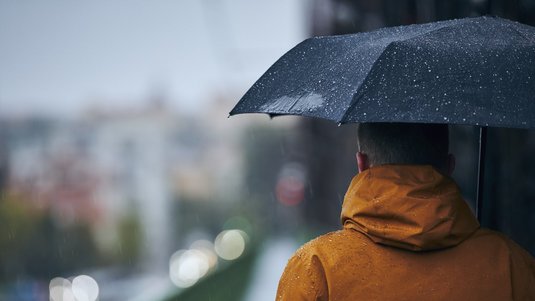 Auf dem Foto ist ein Mann mit einem aufgespannten Schirm von hinten zu sehen, der bei Regenwetter über eine Brücke läuft.