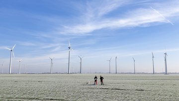 Zwei Personen stehen auf einem Feld umgeben von mehreren Windkraftanlagen unter blauem Himmel