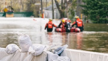 Im Vordergrund befinden sich mehrere aufeinander gestapelte Sandsäcke. Im Hintergrund ist ein überflutetes Gebiet abgebildet, auf dem ein Rettungsboot schwimmt.