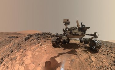 Der Marsrover Curiosity auf dem steinigen, sandigen Marsboden. Das Foto ist aus einem niedrigen Winkel aufgenommen. 