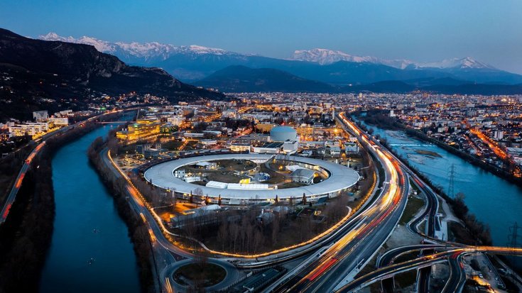 Nachtaufnahme der Synchrotronquelle ESRF in Grenoble | Bildquelle: ESRF/vuedici.org Ein rundes Gebäude zwischen zwei Flüssen in der Dämmerung, von oben fotografiert.
