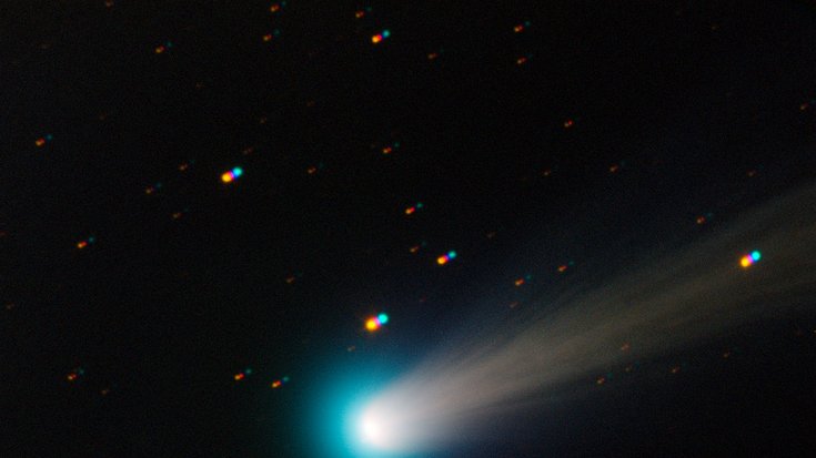 In der Mitte des Bildes ein hell leuchtender Fleck (der Komet), gefolgt von einem schwach leuchtenden Schweif, auf dunklem Hintergrund. Im Hintergrund sind außerdem einige kleine leuchtende Punkte zu sehen (Sterne).