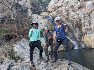 Zwei Frauen und ein Mann stehen mit Werkzeugen in der Hand vor einem kleinen Gewässer, das von einem Wasserfall gespeist wird und in dessen Hintergrund eine Felswand steil aufragt.