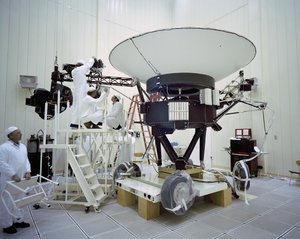 Vier Ingenieure arbeiten an Voyager 2. Die Sonde steht in einer Halle, ihre große Antennenschüssel zeigt nach oben.