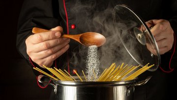 Spaghetti befinden sich in einem Top, über dem sich eine Hand befindet, die von einem Löffel Salz in das Nudelwasser rieseln lässt