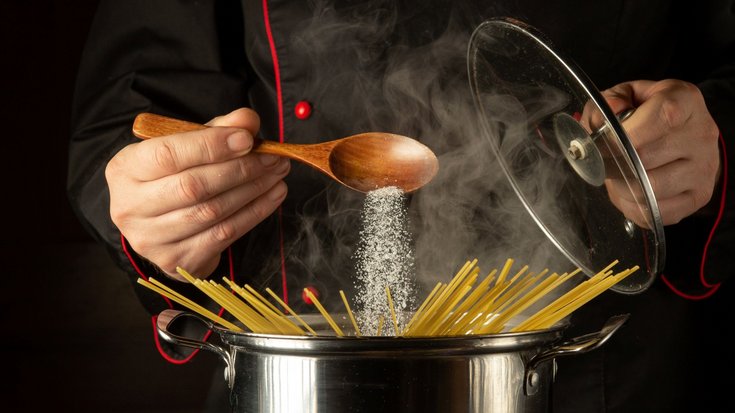 Spaghetti befinden sich in einem Top, über dem sich eine Hand befindet, die von einem Löffel Salz in das Nudelwasser rieseln lässt