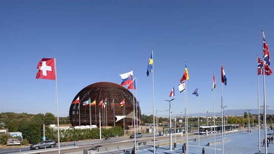 Die Flaggen der Mitgliedstaaten flattern vor dem Kugelhausbau am CERN im Wind. Der Himmel ist wolkenlos.