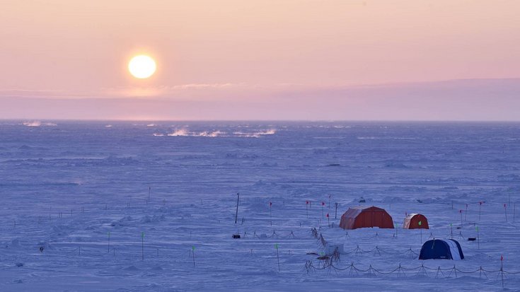 Mehrere Zelte in einer Eislandschaft, am Horizont eine tiefstehende Sonne.